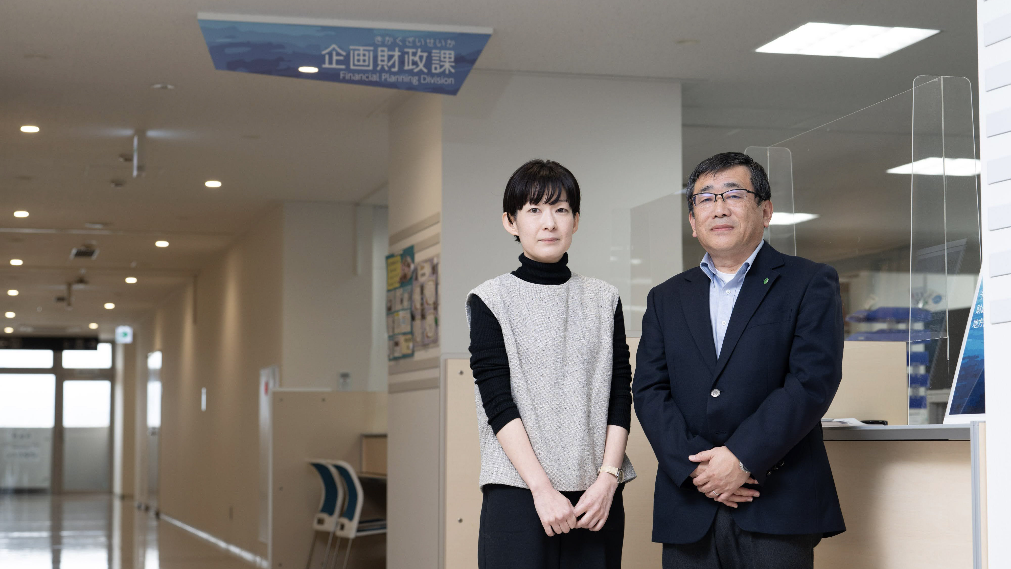 デジタル庁ニュースのサムネイル。築上町の舛川係長（左）と椎野課長が、役所の執務スペースの目に並んで立っている様子を撮影。背後には、天井に「企画財政課」という看板が吊り下げられている。