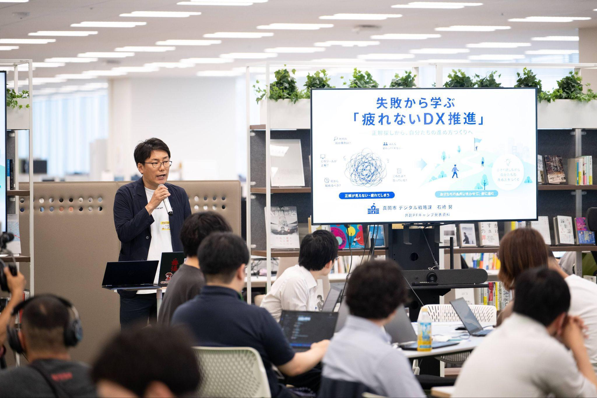 栃木県真岡市の担当者である石崎努氏が右手にマイクを持って、登壇している様子の写真。石崎氏の隣にはモニターが設置されており、「失敗から学ぶ「疲れないDX推進」」と書かれているスライドが映されている。参加者が石崎氏の話を聴きながら、モニターを見ている後ろ姿も写されている。