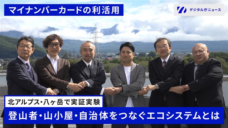 「マイナンバーカードの利活用」というタイトルで、北アルプス・八ヶ岳を背景にスーツ姿の6名の男性が横一列に並んで立っている写真。画面上部に青い帯で「マイナンバーカードの利活用」、右上に「デジタル庁ニュース」のロゴ。画面下部には青い帯で「北アルプス・八ヶ岳で実証実験」「登山者・山小屋・自治体をつなぐエコシステムとは」というテキストが表示されている。背景には雲がかかった山々と青空、緑の山肌が広がり、遠くに街並みが見える。