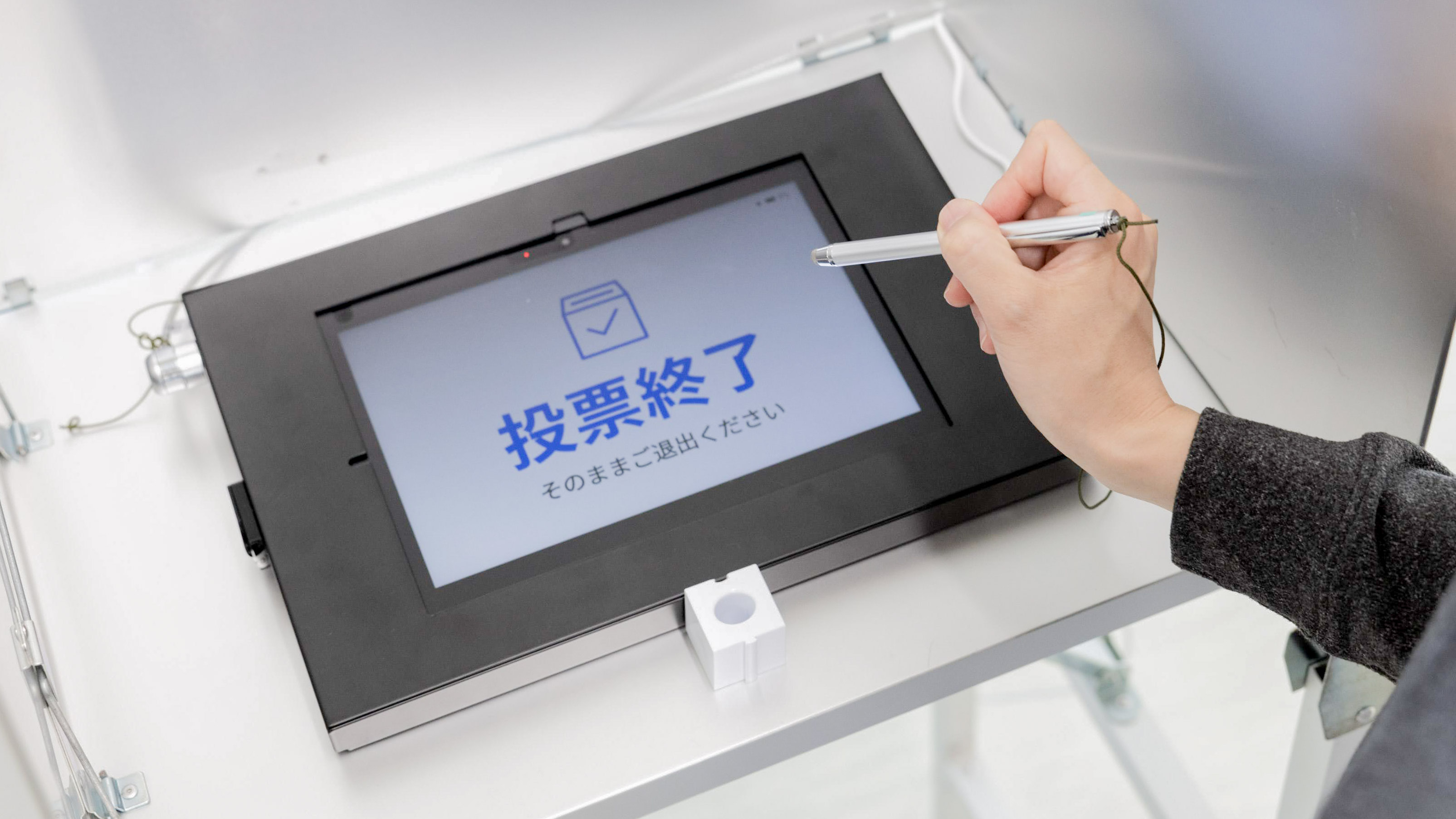 仕切りで囲まれた記載台に、黒いフレームのタブレット型電子投票端末が設置されている。画面には「【投票を開始】を押してください」のメッセージと投票箱のアイコン、青いボタン「投票を開始」が表示されている。スタイラスペンを持ち、画面を操作しようとしている手元が写っている。端末の手前にはペン立てが置かれている。背景の上部には候補者名が書かれた掲示の一部が見える。