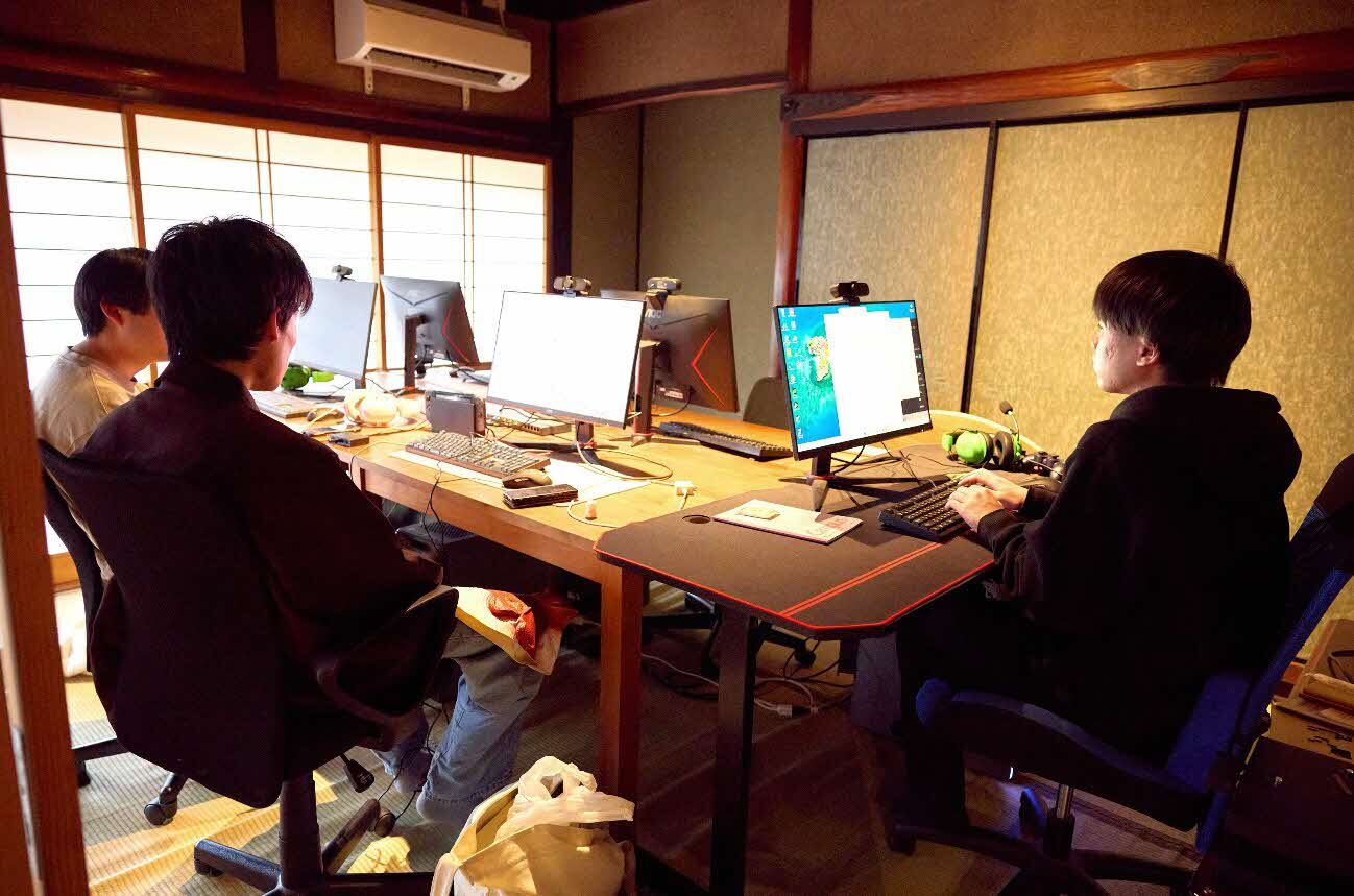 築100年超の古民家を改修して生まれた島根県江津市にあるデジタル拠点「つのづBase」の一室。障子や土壁、木製の梁が残る和室を改装したゲーミングスペースで、3人の若い男性（いずれも後ろ姿）が椅子に座り、デスクに並んだ複数のPCモニターに向かっている。デスク上にはゲーミングキーボード、ウェブカメラ、ヘッドセットなどの周辺機器が置かれており、伝統的な和の空間とハイテク機器が混在した独特の雰囲気を持つ。