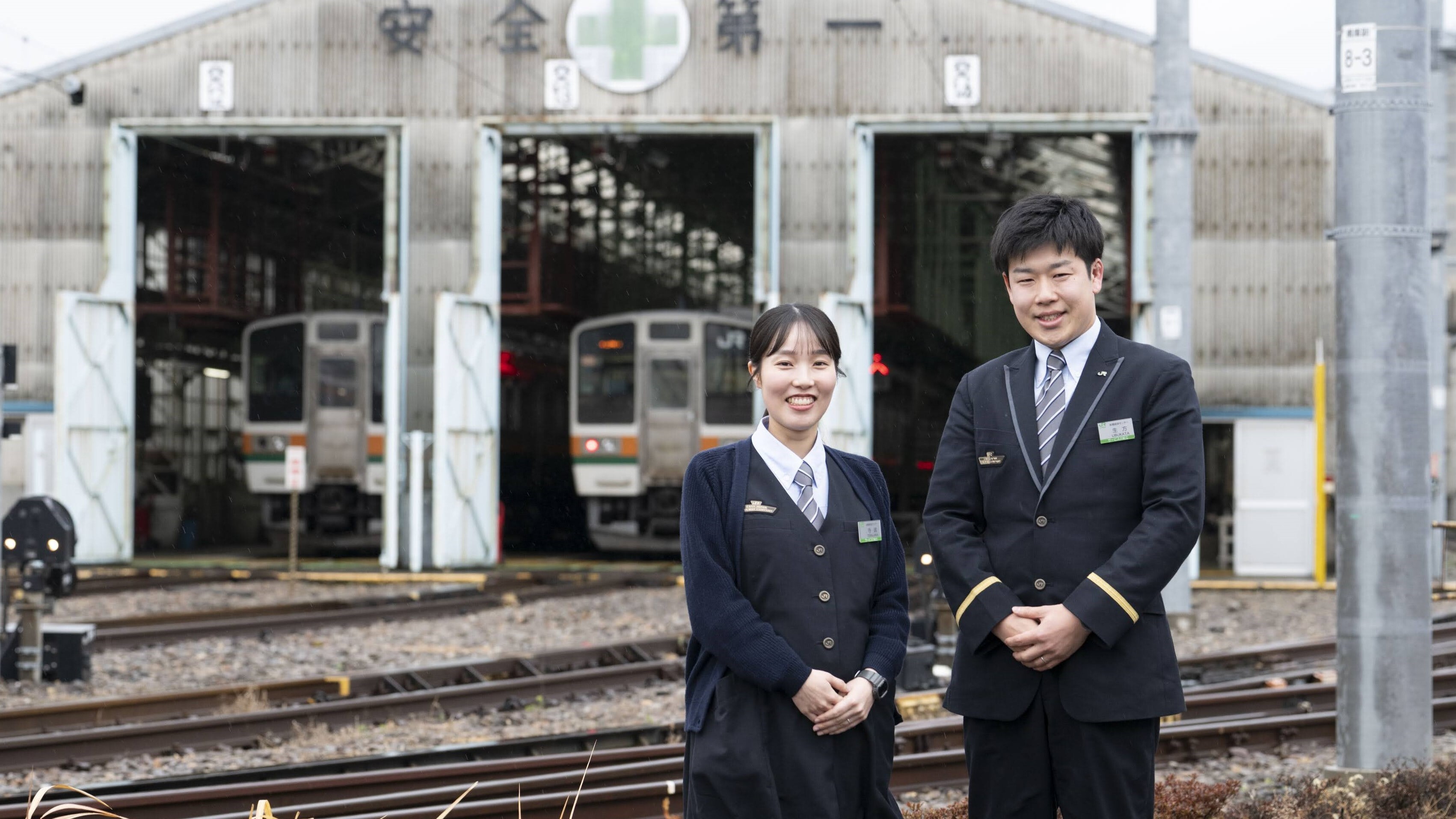 JR東日本の制服を着用した前橋統括センター乗務ユニットの寺嶋彩乃氏（左）と生方聡氏（右）が、高崎車両センターの車両格納庫前を背景に立っている。背後の格納庫には「安全第一」の文字と緑十字のマークが掲げられており、格納庫内にはJR東日本の列車が停車している。
