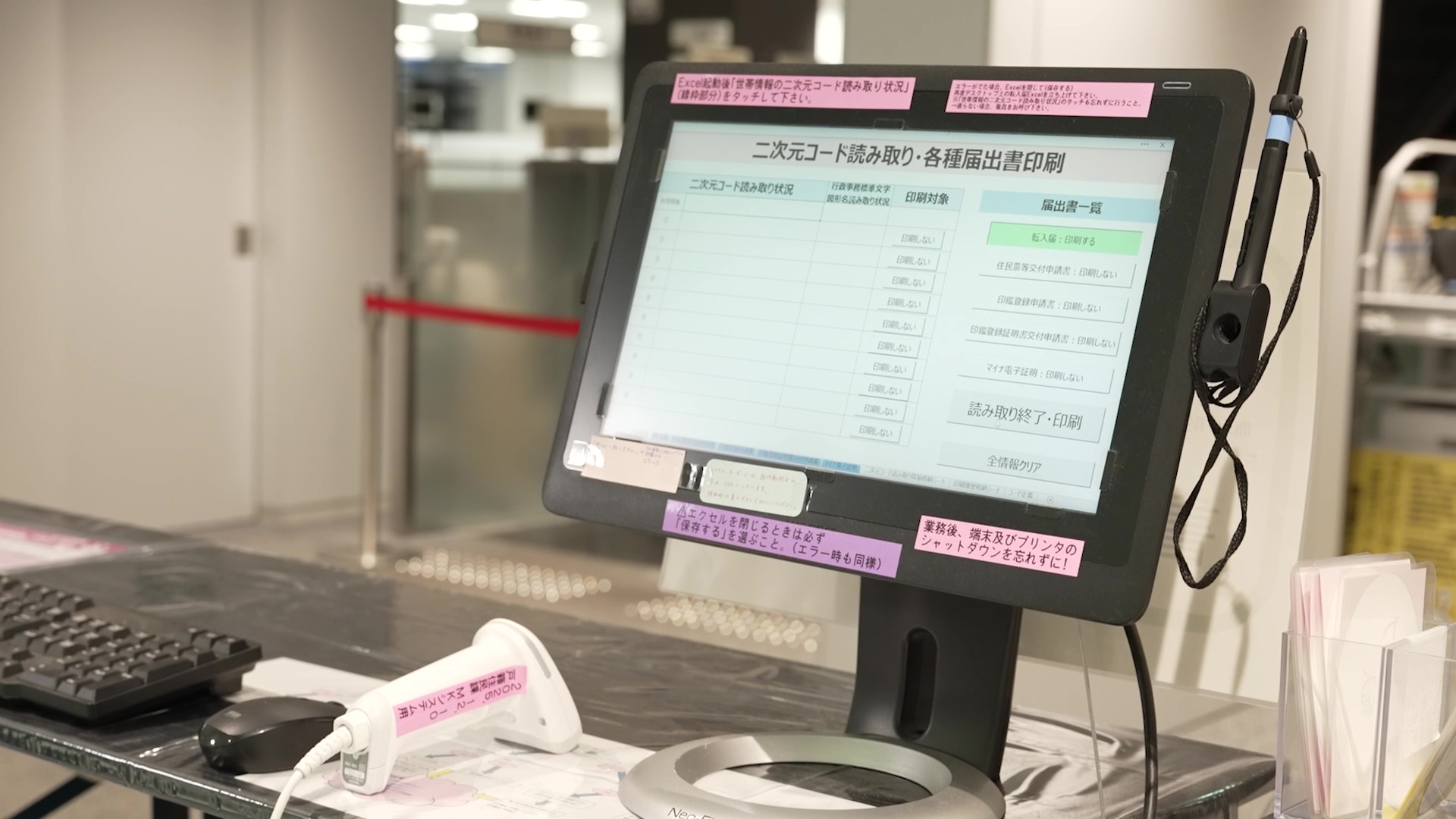 デジタル庁ニュースのサムネイル画像。中の区役所窓口に設置されたタッチパネル式のディスプレイ端末（スタンド型）を正面やや斜めから撮影した写真。画面には「二次元コード読み取り・各種届出書印刷」システムが表示されており、届出書一覧の「転入届：印刷する」が緑色で強調されている。端末の前にはバーコードスキャナー（白色）、キーボード、マウスが置かれている。モニターの四辺にはピンク・紫・黄色の付箋紙で「Excelを閉じるときは必ず『保存する』を選ぶこと。（エラー時も同様）」「業務後、端末及びプリンタのシャットダウンを忘れずに！」などの業務注意メモが貼られている。