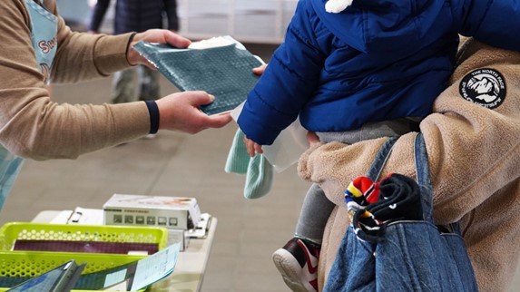 仮受付のシーン。受付台でベージュの上着を着たスタッフの手が、青いバインダーを差し出している。受け取る側は青いジャンパーを着た乳幼児を抱っこしており、子どもはグレーの靴下と運動靴を履いている。手前には緑のトレーや書類・文具類が置かれている。