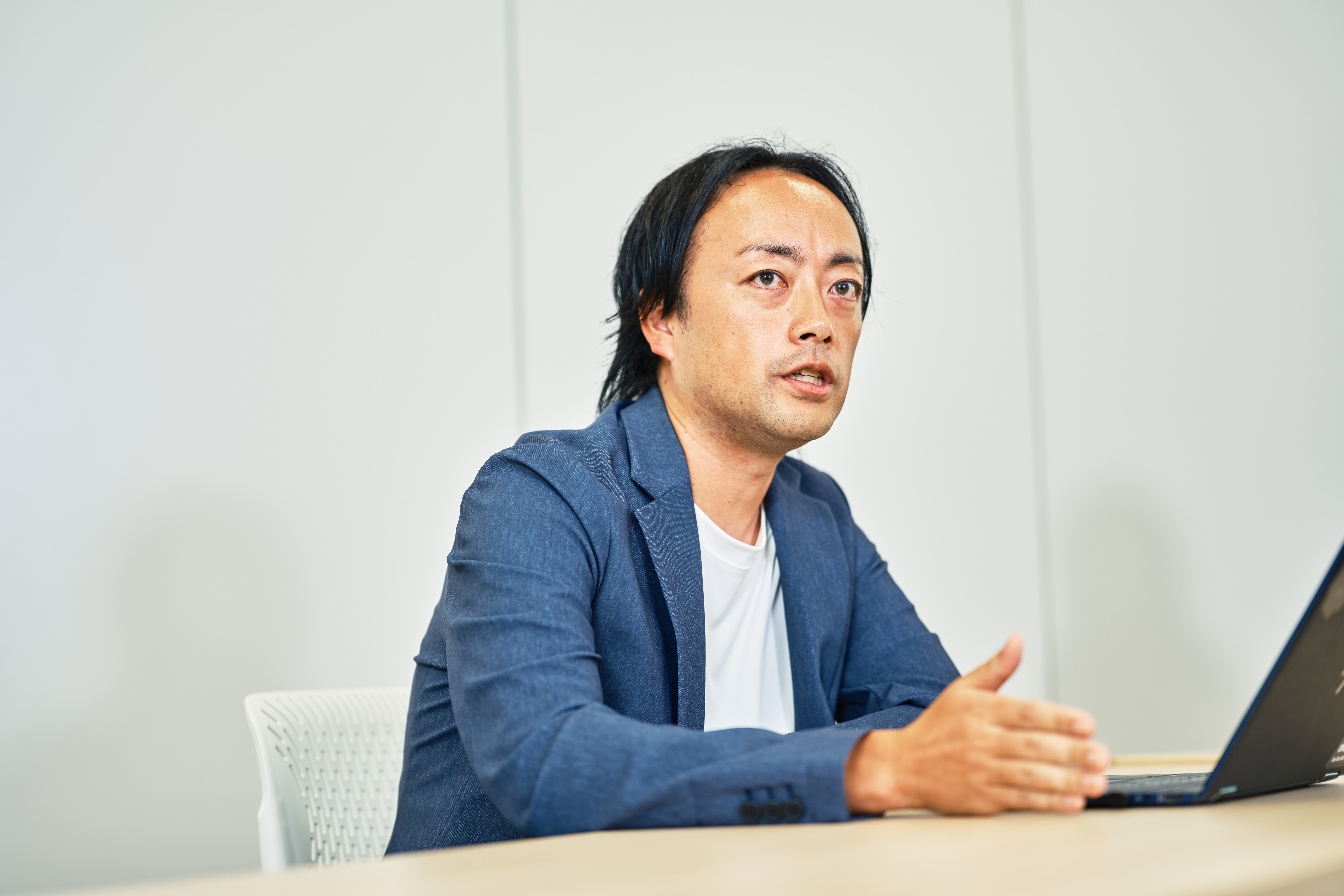 プロダクトオーナーの青野が、会議室の壁を背景に着席して話す様子を向かって左手から撮影した写真。